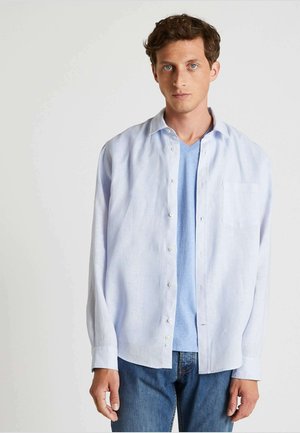 Chemise boutonnée en lin bleu clair à manches longues, dotée d'une poche frontale et d'une texture légère, superposée à un t-shirt V-neck bleu clair.