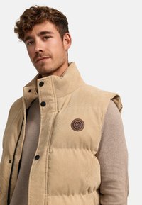 Beige Cordjacke mit Stehkragen, schwarzen Druckknöpfen und einem runden Logoakzent. Über einem hellbraunen Pullover getragen.