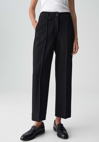 Donna che indossa pantaloni neri sartoriali con pieghe frontali, tasche laterali, bottone e passanti per cintura, abbinati a mocassini neri e a un top bianco senza maniche.