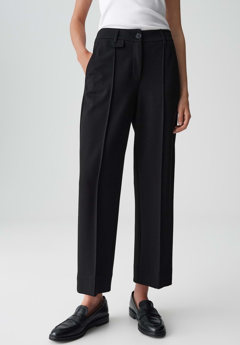 Donna che indossa pantaloni neri sartoriali con pieghe frontali, tasche laterali, bottone e passanti per cintura, abbinati a mocassini neri e a un top bianco senza maniche.