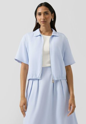 Femme aux longs cheveux noirs portant une veste zippée à manches courtes bleu clair, une jupe plissée assortie, un haut blanc et des boucles d'oreilles argentées en forme d'anneau.