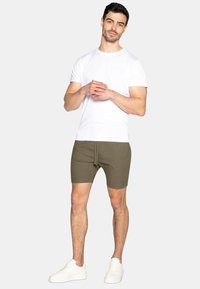 Weißes T-Shirt mit kurzen Ärmeln und Rundhalsausschnitt, kombiniert mit olivgrünen Shorts mit Kordelzug. Modell trägt weiße Sneaker und steht entspannt da.