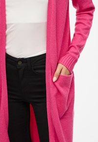 Lange roze cardigan gemaakt van zachte stof met geribde manchetten. Heeft zijzakken en een losse pasvorm, gecombineerd met zwarte jeans en een wit topje.