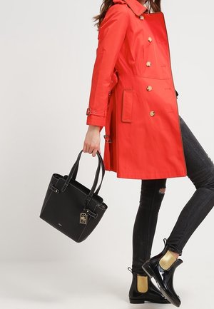 Frau trägt leuchtend roten, doppelreihigen Trenchcoat, schwarze, zerrissene Skinny Jeans, schwarze Lack-Chelsea-Boots mit goldenen Elastikpaneelen und hält eine schwarze Handtasche.