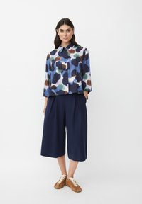 Donna in piedi con le mani nelle tasche dei pantaloni a palazzo blu scuro, indossa una camicetta a righe multicolore e sneaker marroni.