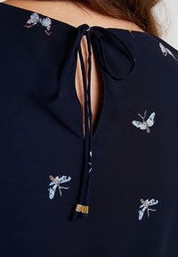 Námořnická blůza s klíčovou průsvitnou zádovou částí a stuhou na zavázání, zdobená potiskem světle modrých a růžových motýlů. Hladký textilní povrch.