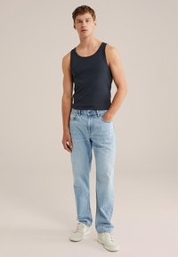 Joven de pie con una mano en el bolsillo, vistiendo una camiseta sin mangas negra, jeans azules claros y zapatillas blancas sobre un fondo simple.