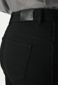 Czarne dżinsy o gładkiej fakturze, z skórzanym naszywką na pasku, tylnymi kieszeniami oraz kontrastowymi detalami stębnowania.