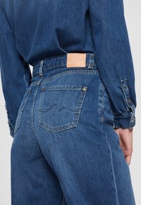 Personne portant un jean bleu taille haute et une chemise en jean bleue assortie, vue de dos, avec un patch en cuir au-dessus de la poche.