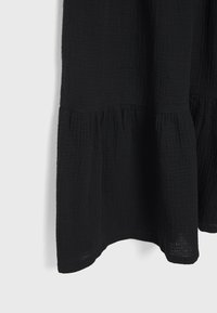 Robe en tissu noir texturé avec un volant froncé près de l'ourlet, suspendue sur un fond blanc uni.