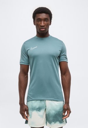 Homme debout portant un t-shirt Nike à manches courtes couleur sarcelle et un short de sport à motif nuages sur fond uni.