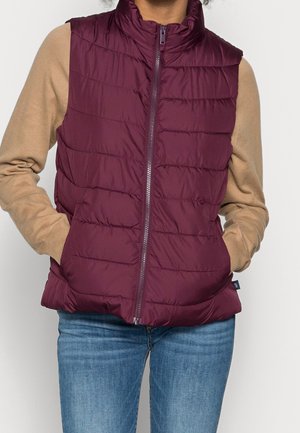 Personne portant un gilet matelassé bordeaux avec une fermeture éclair à l’avant sur un t-shirt beige à manches longues et un jean bleu, mains dans les poches du gilet.