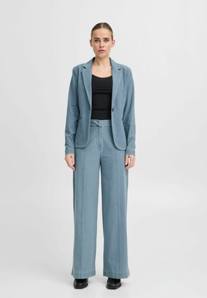 Hellblauer maßgeschneiderter Blazer und weit geschnittene Hose, ausgestattet mit einer weichen Textur und Knopfdetails, kombiniert mit einem schwarzen enganliegenden Oberteil.