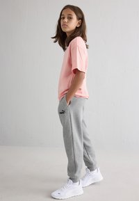 Gaiši rozā īsām piedurknēm T-krekls, pelēki sporta bikses ar logotipu un baltas sporta apavus. Modelis ar taisniem brūniem matiem, stāv sānu profilā.