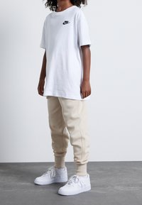 Bílý bavlněný tričko s černým logem Nike, doplněné béžovými tepláky a bílými teniskami. Měkké materiály a uvolněný střih jsou v designu jasně patrné.