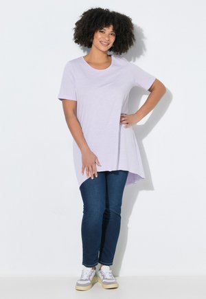 Uśmiechnięta kobieta o kręconych włosach, ubrana w jasnofioletową asymetryczną tunikę, ciemnoniebieskie dżinsy i białe trampki, stojąca na tle białej ściany.