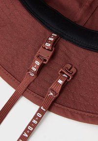 Brown fabric bucket hat features a textured surface, an adjustable strap with a buckle, and printed accents on the strap.
