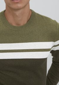 Zeleni pulover s teksturiranim završetkom, s horizontalnim bijelim prugama i okruglim izrezom. Tkanina izgleda meko i pleteno.