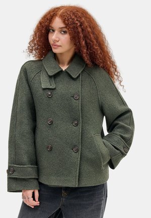 Giovane donna con capelli rossi ricci che indossa un cappotto di lana verde scuro doppiopetto con grandi bottoni e tasche laterali.