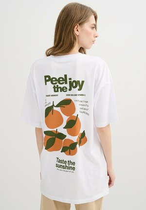 Mujer con cabello castaño claro que lleva una camiseta blanca con una ilustración de una naranja y el texto "Pela la alegría" y "Prueba el sol" en la parte trasera.