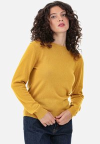 Senfgelber Strickpullover mit Rundhalsausschnitt, gerippten Bündchen und Saum sowie langen Ärmeln; getragen über dunklen Denim-Hosen.