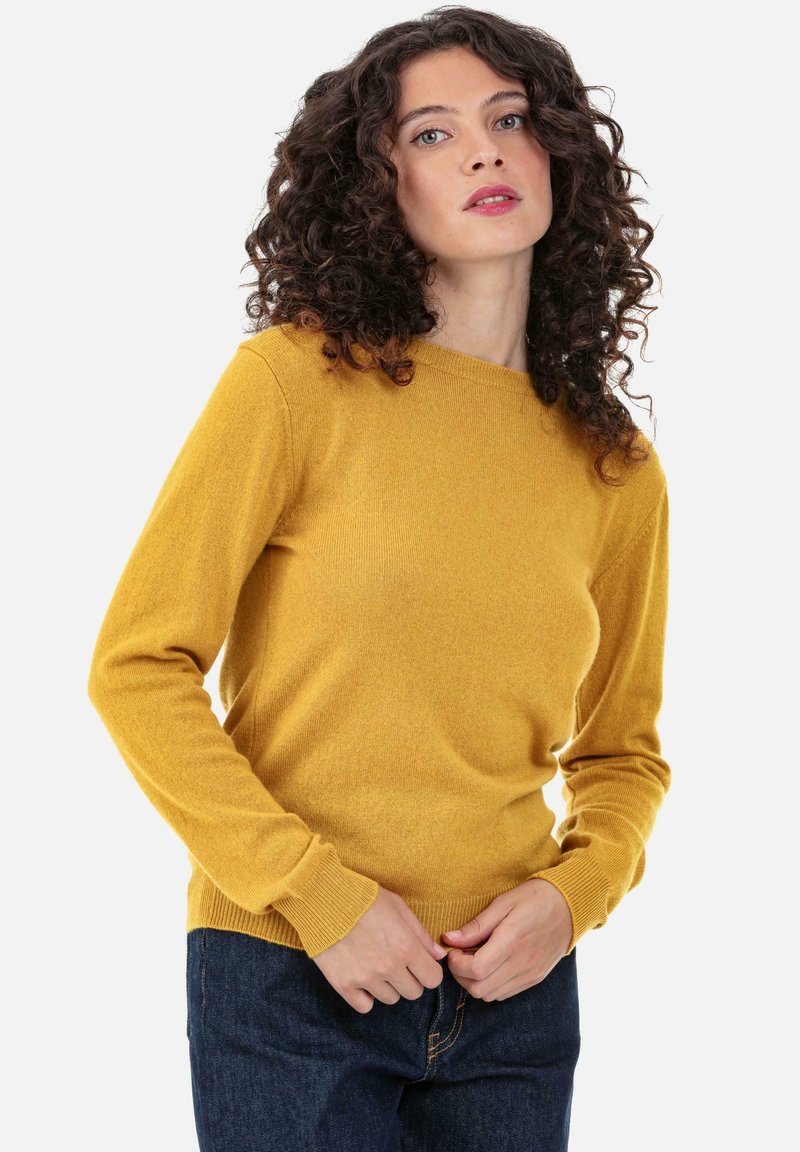 Senfgelber Strickpullover mit Rundhalsausschnitt, gerippten Bündchen und Saum sowie langen Ärmeln; getragen über dunklen Denim-Hosen.