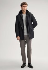 Uomo che indossa un cappotto grigio scuro sopra una giacca trapuntata nera, un maglione in misto lana grigio chiaro, pantaloni marroni e stivali in pelle nera, in piedi contro uno sfondo semplice.