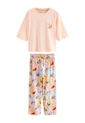 Kvinde stående på sengen med armene hævet iført en lyserød oversized top og farverige blomstrede pyjamasbukser, smilende med lukkede øjne.