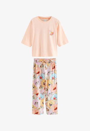Kvinde stående på sengen med armene hævet iført en lyserød oversized top og farverige blomstrede pyjamasbukser, smilende med lukkede øjne.