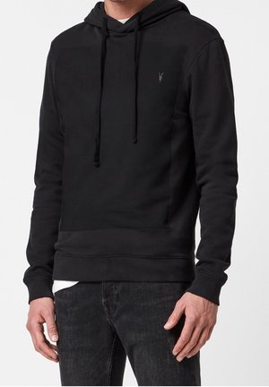 Mand iført en enkel sort hættetrøje med snore og en lille logo på brystet, kombineret med mørke jeans, stående mod en ensfarvet baggrund.