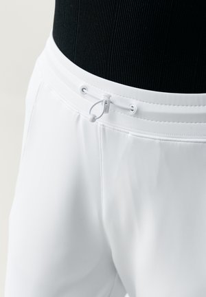  bílé kalhoty s elastickým pasem, vybavené bílým nastavitelným tkaničkovým páskem a sponou, nošené s černým žebrovaným topem.