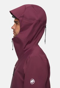 Burgunderfarbene wasserdichte Jacke mit großer Kapuze, seitliche Ansicht. Mit einem Logo auf dem Ärmel und verstellbaren Kordeln an der Kapuze.