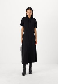 Schwarzes Midi-Kleid aus leichtem Stoff mit kurzen Ärmeln, hohem Kragen und einem Schlitz vorne. Kombiniert mit kniehohen schwarzen Stiefeln und einer silbernen Tasche.