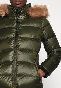 Olivgrön vadderad jacka med glänsande yta, dragkedja, sidofickor och avtagbar kant av fuskpäls på huvan.