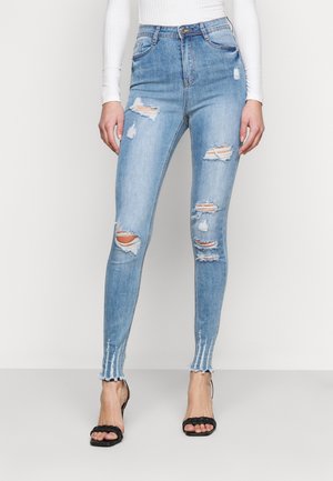 Femme portant un jean skinny déchiré taille haute bleu clair et des sandales noires à talons, debout devant un fond uni.