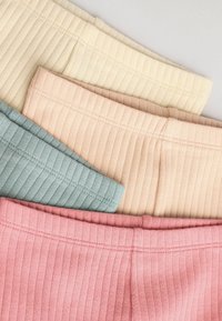 Quatre vêtements en tissu côtelé pliés dans des couleurs pastel crème, beige, bleu clair et rose disposés en pile diagonale.