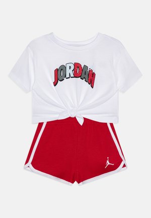 Weißes Crop-Top mit "JORDAN" in kräftigen Farben, kombiniert mit roten Shorts mit weißen Seitenstreifen und einem kleinen weißen Logo am Saum.