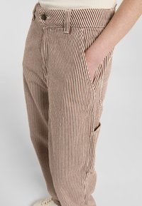 Gestreifte Hose in Beige und Braun, mit hohem Bund, Seitentaschen und lässigem Schnitt. Der Stoff hat eine strukturierte Oberfläche.