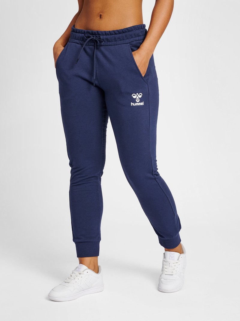 Vrouw die marineblauwe hummel joggers draagt met een trekkoord in de taille en witte sneakers, staand met handen in de zakken tegen een witte achtergrond.