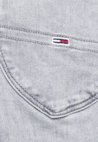 Tejido de mezclilla gris con una costura de bolsillo visible. Presenta un pequeño logotipo bordado con rayas rojas y azules sobre un fondo blanco.