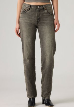 Kvinde iført grå jeans med høj talje og lige ben og sorte ankelstøvler, stående foran en ensfarvet lys baggrund.