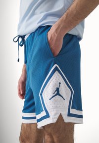 Siniset verkkokankaiset urheilushortsit, joissa on timantinkaltainen valkoinen yksityiskohta ja musta Jumpman-logo. Sisältää sivutaskut ja narukuuluksen vyötäröllä.
