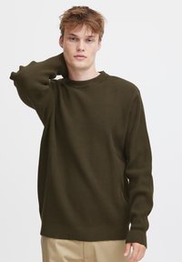 Olivenfarget strikkegenser med teksturert mønster, rund hals og lange ermer. Ribbestrikkede mansjetter og nederkant gir en tettsittende finish.