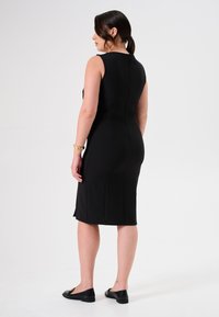 Schwarzes ärmelloses Kleid mit tailliertem Schnitt, knielang, mit Nahtdetails und einem kleinen Schlitz hinten. Getragen mit schwarzen Ballerinas.