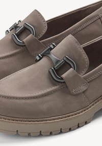 Mocassins en suède gris avec une semelle en caoutchouc épaisse et un détail en métal noir en forme de mors sur le dessus, présentant des coutures visibles autour des bords.