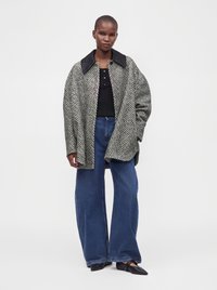 Γυναίκα που φοράει μαύρο-λευκό oversized σακάκι με σχέδιο ψαροκόκαλο, μαύρο τοπ, φαρδιά μπλε τζιν και μαύρα μυτερά παπούτσια, που στέκεται μπροστά σε λευκό φόντο.