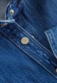 Donkerblauwe denim close-up met een metalen drukknoop en contrasterende stiksels op een kraagdetail. Een gladde textuur en een casual ontwerp.