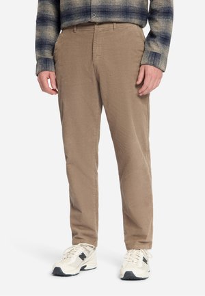 Mann trägt beige Cordhosen, weiße Sneakers und ein blau-grau kariertes Langarmhemd, steht vor weißem Hintergrund.