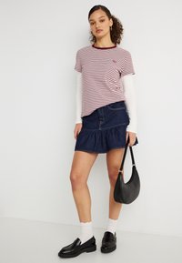 Γυναίκα που φορά ριγέ μπλούζα πάνω από λευκά μακρυμάνικα, τζιν φούστα, λευκές κάλτσες, μαύρα μοκασίνια, κρατώντας μαύρη τσάντα απέναντι σε λευκό τοίχο.