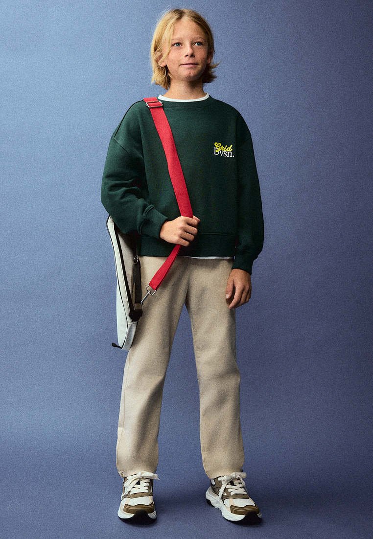 Enfant aux cheveux blonds portant un sweat-shirt vert foncé, un pantalon beige et des baskets, tenant un sac blanc avec une sangle rouge sur un fond uni.